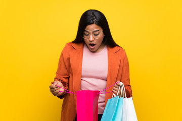 Young Colombian girl over yellow wall surprised while holding a lot of shopping bags