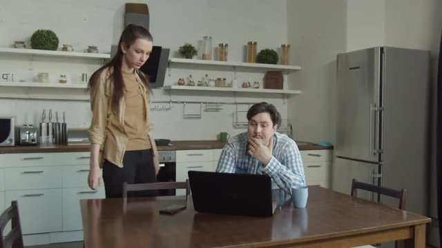 Surprised woman seeing confused husband sitting at kitchen table, chatting online with mistress on laptop. Angry female caught ashamed man viewing embarrassing content online, requiring explanations.