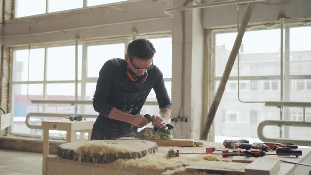 Panning shot of skilled carpenter sanding wooden plank with electric sanding machine, sawdust flying around
