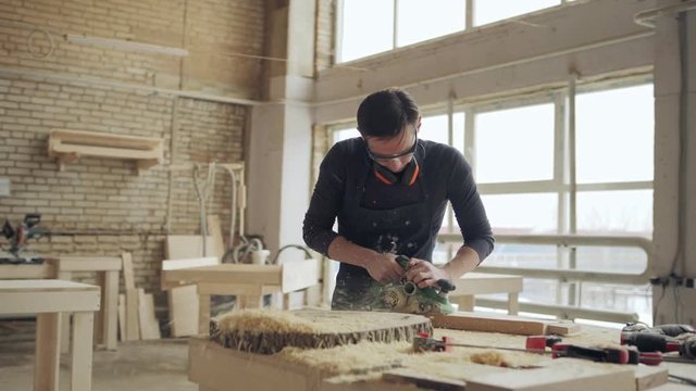 Panning shot of skilled carpenter working in his woodworking workshop. Craftsman sanding wooden plank with electric flatbed sander, sawdust flying around