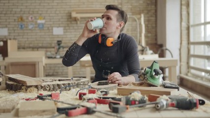 Tracking shot of professional joiner relaxing at workplace. Woodworker having coffee and surfing Internet on smartphone surrounded by tools and wood shavings - Powered by Adobe