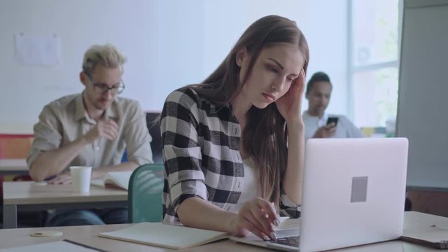 College student girl sitting at desk and using laptop to do difficult research. She making notes in notebook while her male mates studying and using phone in background