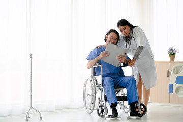 Female doctor take care of smiling asian male patient who sitting on wheelchair  with document file folder. The young man recover from illness. Healthcare and medical concept.