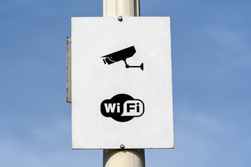 Generic camera surveillance and Wi-Fi signs on a white panel on a street lightning pole towards...