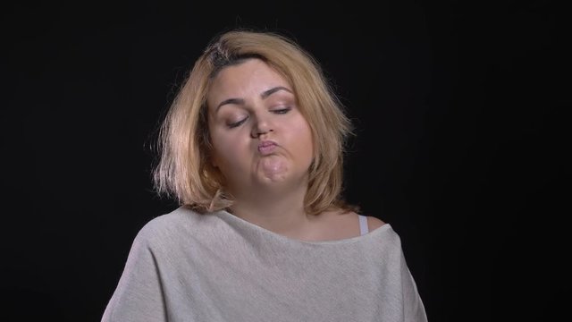Bored blonde short-haired overweight businesswoman blowing up cheeks watching with annoyance on black background.