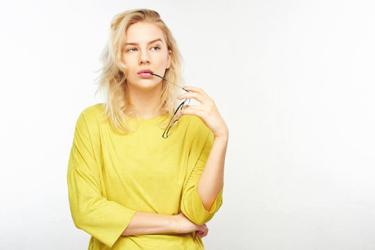 Pensive European stylish young woman in yellow clothes holding glasses near mouth, doubts what to choose, thinks, decides what to buy on white background