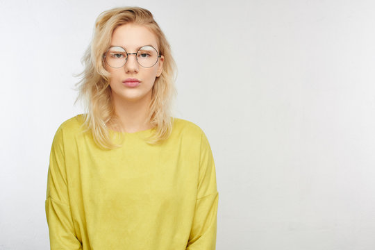 Serious European girl student in round glasses, yellow clothes looking into the camera, worried about entrance exams on a white background with copy space