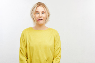 A crazy blond girl in yellow clothes on white background squints eyes, sticks out tongue, makes...