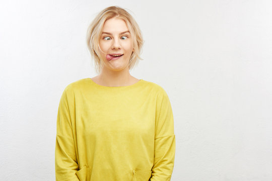A crazy blond girl in yellow clothes on white background squints eyes, sticks out tongue, makes funny fool face, does not want to grow up and be responsible