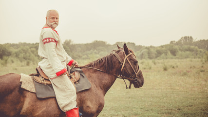 Man in ethnic clothing is riding a horse on the summer fields background.
