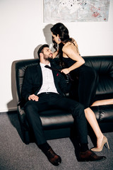 brunette girl in black dress holding tie of passionate man sitting on sofa