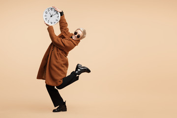 Smiling young hipster woman with blonde short hair wearing a coat and sunglasses dance with clock isolated over beige background.
