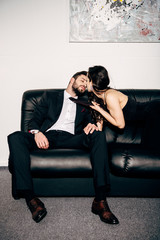 brunette sexy girl in black dress holding tie of passionate man sitting on sofa