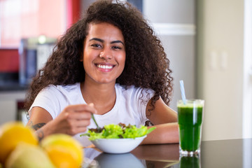 cheerful young afro american woman eating vegetable salad and green juice in home kitchen.