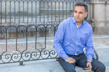 An elegant man in blue shirt waits sitting on a bench in the middle of a street