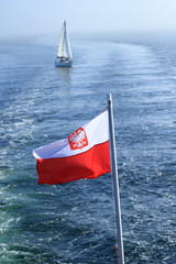 Polish flag waving on the mast of a ship at sea with a yacht in the background
