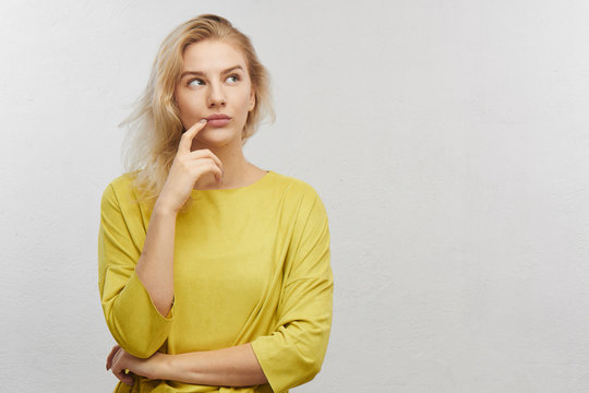 Portrait of embarrassed young woman looks aside holds chin with confused mouth in yellow clothes on white background with free space for your text