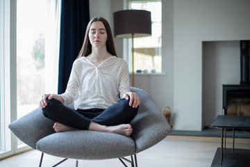 Peaceful Teenage Girl Meditating Sitting In Chair At Home - obrazy, fototapety, plakaty