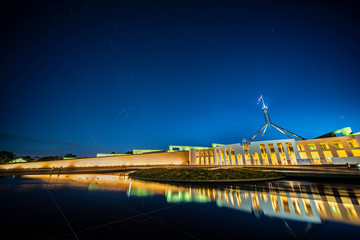 Facade of new parliament house in Canberra on capitol hill at sunset with bright illumination reflecting in blurred waters of pool.  South cross on sky 