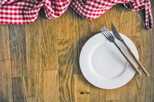 Red checkered dishcloth, white plate and cutlery on wooden background. Culinary background. Table setting. Preparing the food. Top view, copy space