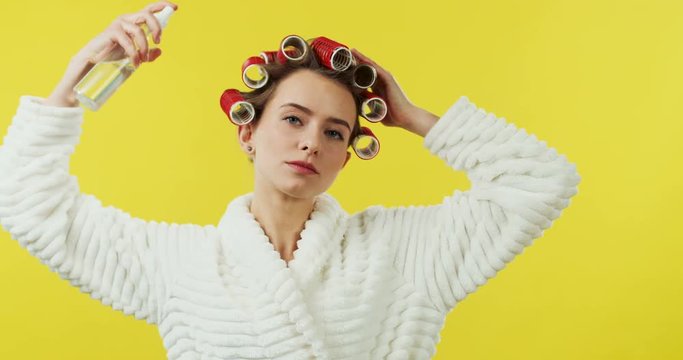 Beautiful Caucasian woman in the white bathrobe and with curlers on the head spraying her heair while using hairspray on the yellow wall back.