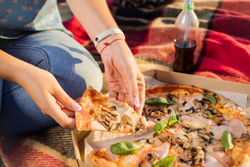 Happy young woman holding hot pizza in box, seat outdoor in park