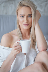 Beautiful blond woman drinking a coffee while sitting on her bed at home