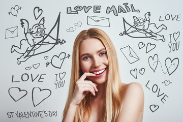 Flirty mood. Young female with naked shoulders touching her lips and looking at camera while standing against grey background with doodles on the theme of St.Valentine's day.