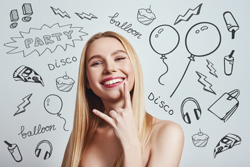 Feeling free. Cute blonde young woman gesturing and smiling at camera while standing against grey background with party theme doodles on it. Having fun.