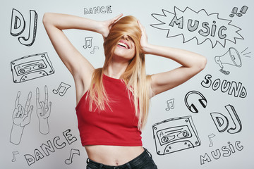 Feeling playful and happy. Slim woman in red tank top covering face with her hair and smiling while standing against grey background with music theme doodles.