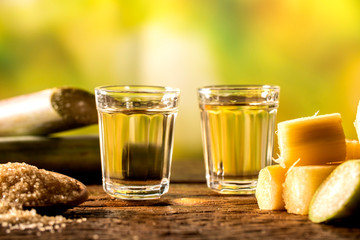 two shot glasses of Brazilian gold  cachaca with sugar and sugarcane isolated on rustic wooden background - obrazy, fototapety, plakaty