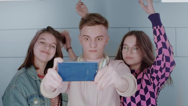 Three teenage friends in bright clothes take selfies with a smartphone. Girls and guy make funny faces, smile, laugh, fool around, correct hairstyles. Close-up portrait. Slow motion. 4K