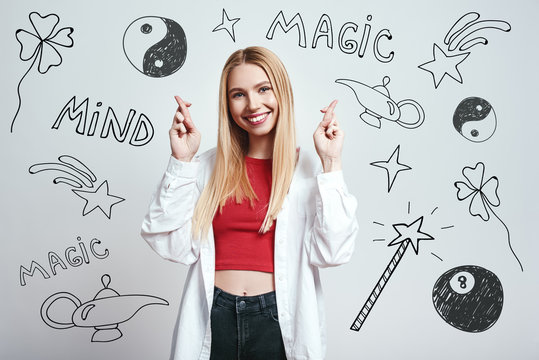 I know magic trick. Smiling young blonde woman in white shirt holding fingers crossed while standing against grey background with hand drawn magic tools on it.