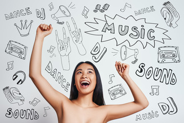 Dancing beauty. Happy asian woman is raising hands up and smiling while standing against grey background with music theme doodles on it.