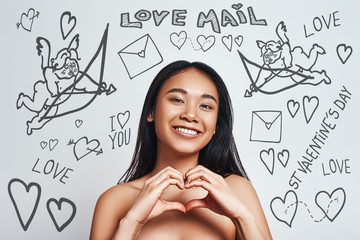 Sending my love. Smiling cute asian woman making a heart shape with her hands while standing against grey background with doodles on the theme of St.Valentine's day.