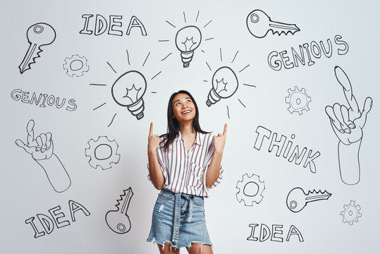 Full of ideas! Cute asian woman with long hair pointing up at pointing at drawn light bulbs above her head and smiling while standing against grey background with hand drawn doodles on it