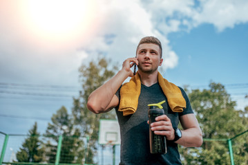 A male athlete, summer city after a workout, calls on phone, holds in his hand a shaker, a bottle of water, a towel around his neck. Resting after a hard workout. Active lifestyle, workout in nature.