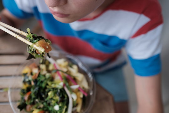 Young boy is eatinh healthy food poke bowl with chinese chopsticks.