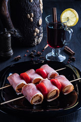 Cheese wrapped in bacon on a stick for kebabs, lies on a square dark plate and a glass of mulled wine with lemon and spices