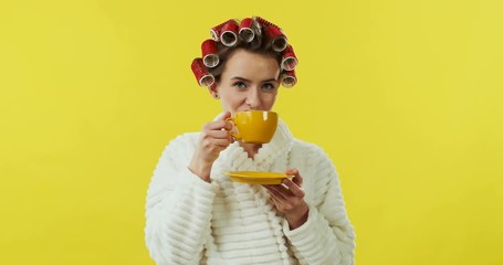 Cheerful young Caucasian pretty girl in curlers and bathrobe laughing to the camera and sipping her drink from the yellow cup.