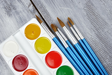 A palette of watercolor paints and brushes of different sizes, lying on a gray wooden background