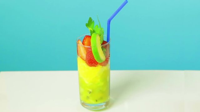 Fruit cocktail in a glass of kiwi, apple and strawberry slices decorated with lime with a blue straw on a blue background. Glass rotates in a circle.