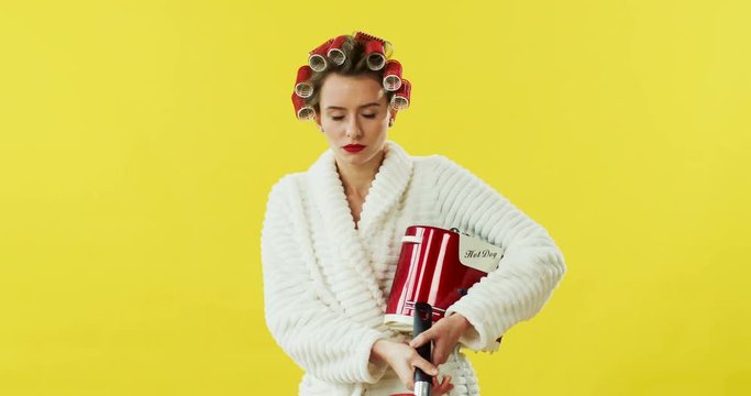 Caucasian angry beautiful young wife in the bathrobe and curlers standing on the yellow background with a hot-dog maker machine and frying pan, waving with it like beating and expressing her