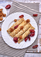 Pancakes, Walnut, Plums. Pancakes with filling on a white plate. Wood background. Rustic style. Rolled up pancakes. Wrapped thin pancakes. Homemade crepes, dried fruits, nuts.