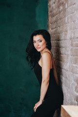 Fototapeta premium Beautiful girl in a black tight-fitting dress and heels. Black long wavy hair. Eastern appearance. Beautiful cut eyes. Brown sofa and green wall. A dark room. Sexy and stingy brunette. Portrait.