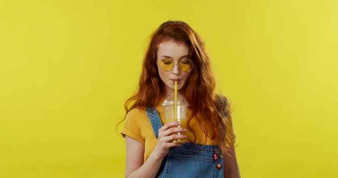 Caucasian young cool stylish girl in sunglasses and with red hair coming in the shot looling flirtly to the camera and going away. Yellow wall on the background.