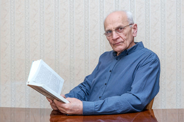 old man with glasses sitting at the table and reading a book, side view