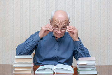 an old man wearing glasses is sitting at a table reading a book, many books are stacked around