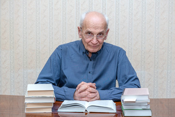 old man sits at a table and looks out from under his glasses, a stack of books around