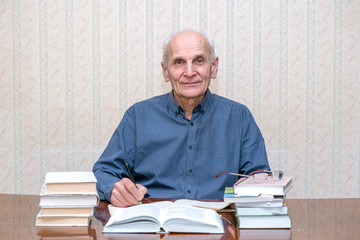 mature professor with a ballpoint pen in his hands sitting at a table with a lot of books